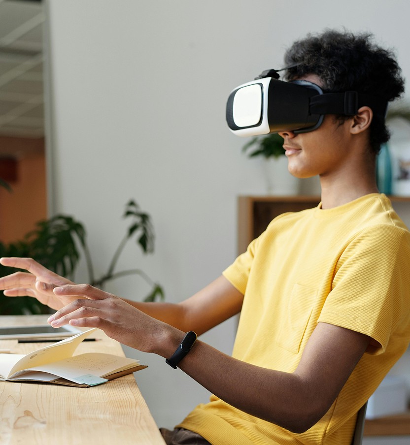 A young person wearing a yellow shirt sits at a desk with a VR headset, gesturing with hands as if interacting with a virtual environment. Relaxed atmosphere.